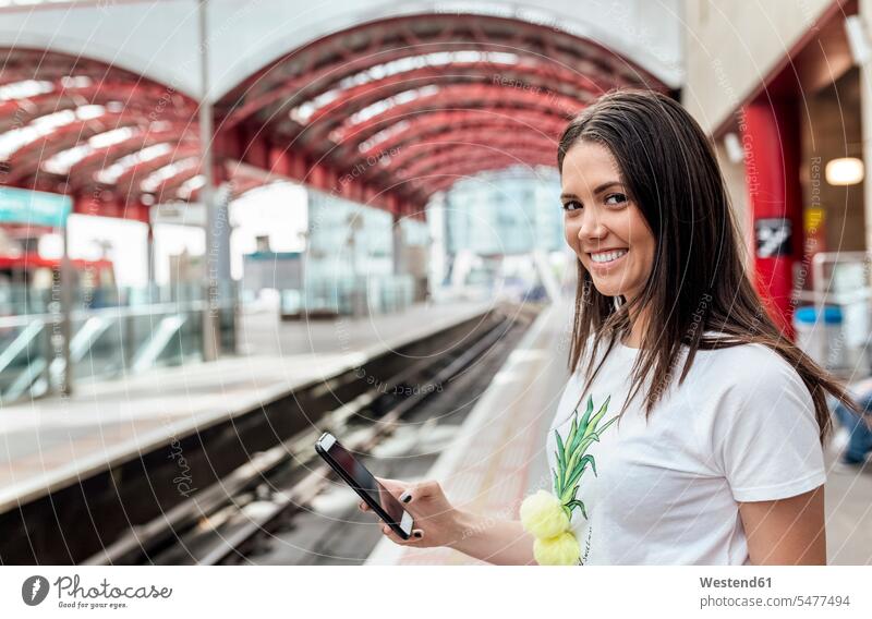 UK, London, portrait of smiling young woman holding cell phone in a train station portraits smile mobile phone mobiles mobile phones Cellphone cell phones