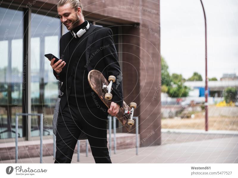 Smiling fashionable young man holding cell phone and skateboard passing office building Businessman Business man Businessmen Business men Skate Board