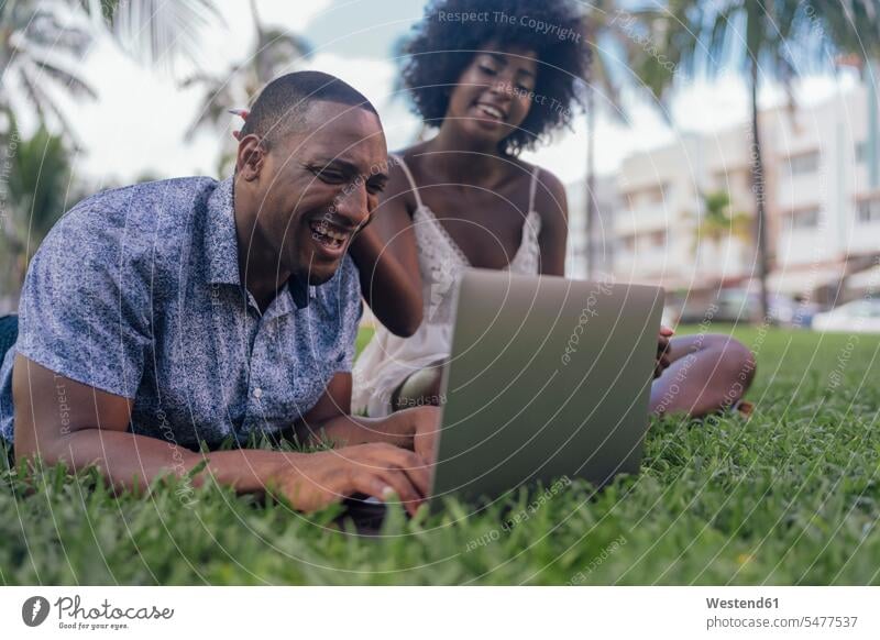USA, Florida, Miami Beach, laughing young couple looking at laptop on lawn in a park eyeing Laptop Computers laptops notebook parks Laughter twosomes
