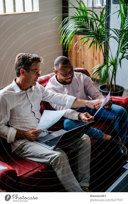 Two businessmen using laptop and discussing documents on sofa in loft office offices office room office rooms discussion couch settee sofas couches settees
