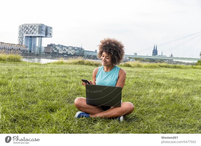 Germany, Cologne, woman sitting on meadow using laptop and cell phone females women Laptop Computers laptops notebook mobile phone mobiles mobile phones