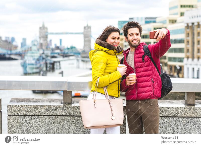 UK, London, young couple with coffee to go standing on bridge over the Thames taking selfie with smartphone twosomes partnership couples Smartphone iPhone