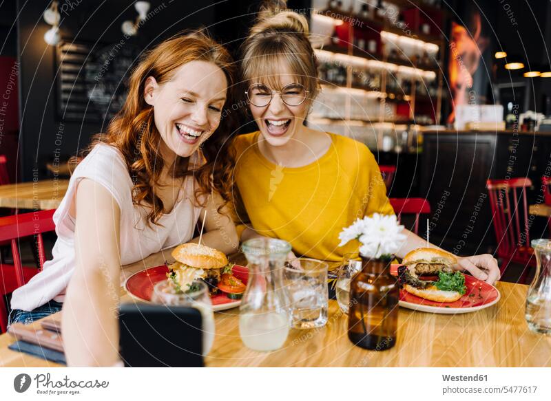 Two happy female friends having burger and taking a selfie in a restaurant Crockery Tableware dish dishes Plates telecommunication phones telephone telephones