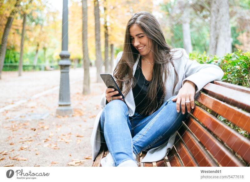 Smiling woman sitting on a park bench using smartphone benches park benches telecommunication phones telephone telephones cell phone cell phones Cellphone