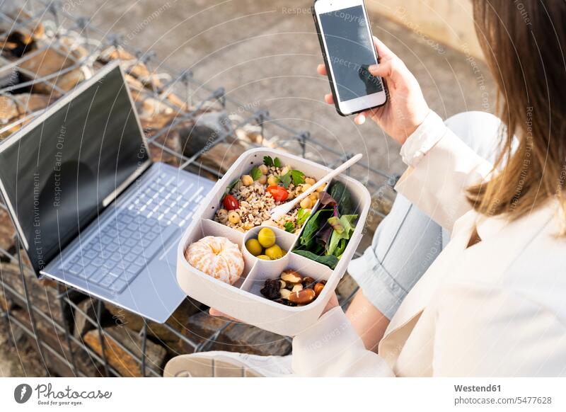 Female entrepreneur using smart phone while holding reusable lunch box color image colour image outdoors location shots outdoor shot outdoor shots day