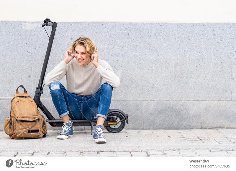 Smiling man with backpack wearing headphones while sitting on electric push scooter against wall color image colour image outdoors location shots outdoor shot