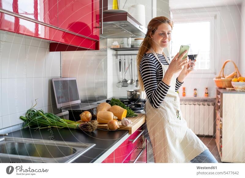 Woman drinking glass of red wine in the kitchen, while using smartphone human human being human beings humans person persons adult grown-up grown-ups grownup