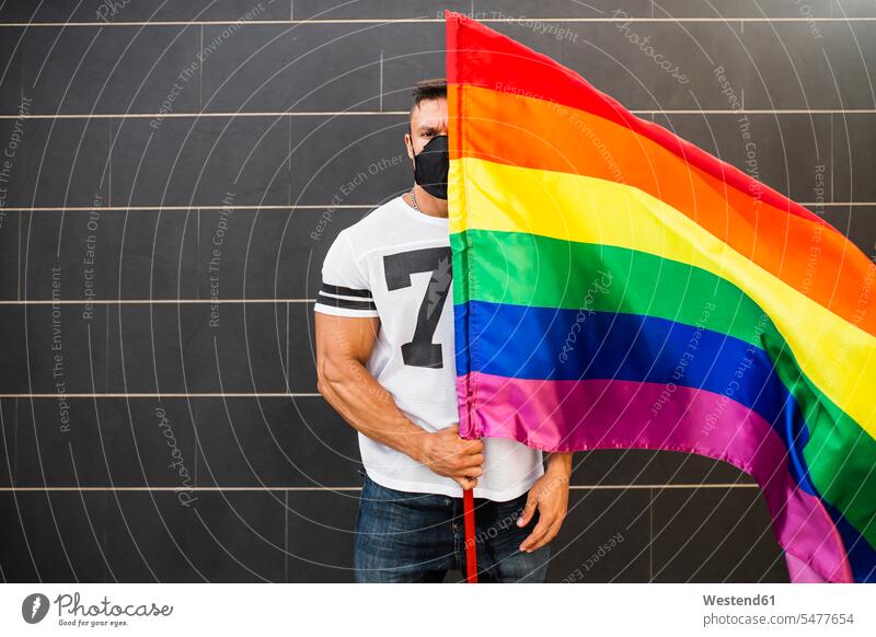 Young man wearing mask holding rainbow flag while standing against wall color image colour image Spain outdoors location shots outdoor shot outdoor shots day
