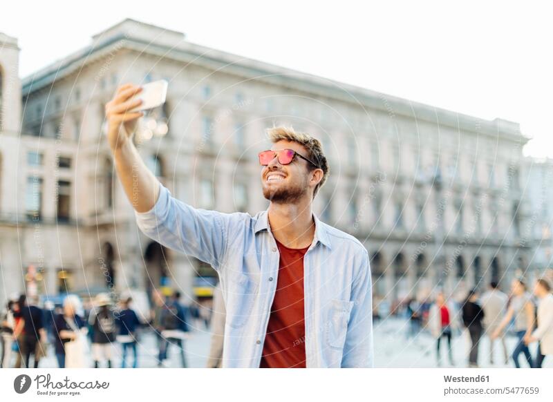 Smiling young man taking a selfie in the city, Milan, Italy touristic tourists shirts telecommunication phones telephone telephones cell phone cell phones