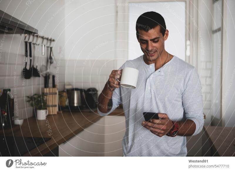 Smiling young man with cup of coffee using cell phone in kitchen at home men males Coffee smiling smile Coffee Cup Coffee Cups mobile phone mobiles