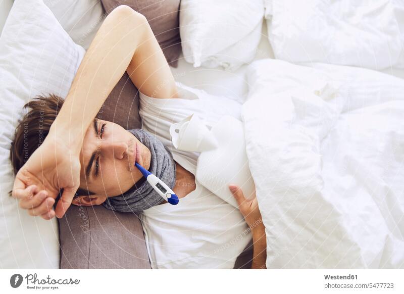 Sick young man with thermometer in mouth resting on bed at home color image colour image Germany indoors indoor shot indoor shots interior interior view