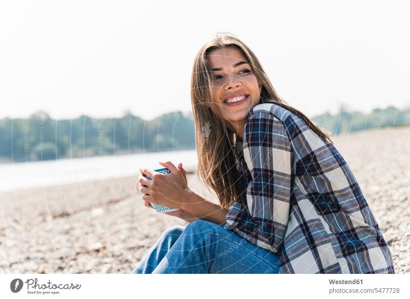 Happy young woman holding a mug sitting at the riverside mugs cup riverbank happiness happy females women Seated water's edge waterside shore Adults grown-ups