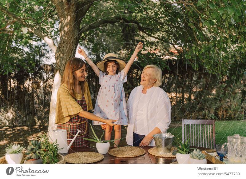 Cheerful girl with arms raised standing with mother and grandmother against tree in yard color image colour image Spain leisure activity leisure activities