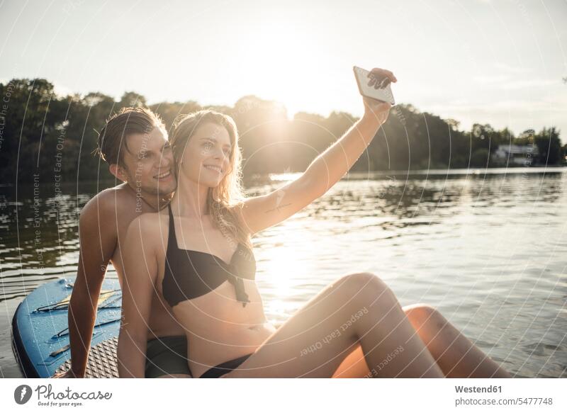 Happy couple sitting on paddleboard on a lake, taking smartphone selfies human human being human beings humans person persons caucasian appearance