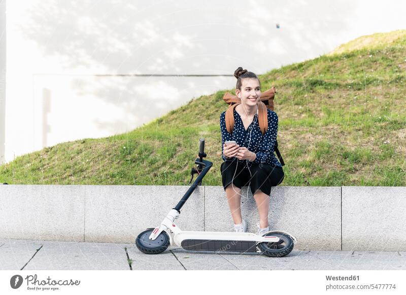 Smiling young woman with electric scooter, earphones and cell phone having a break human human being human beings humans person persons caucasian appearance