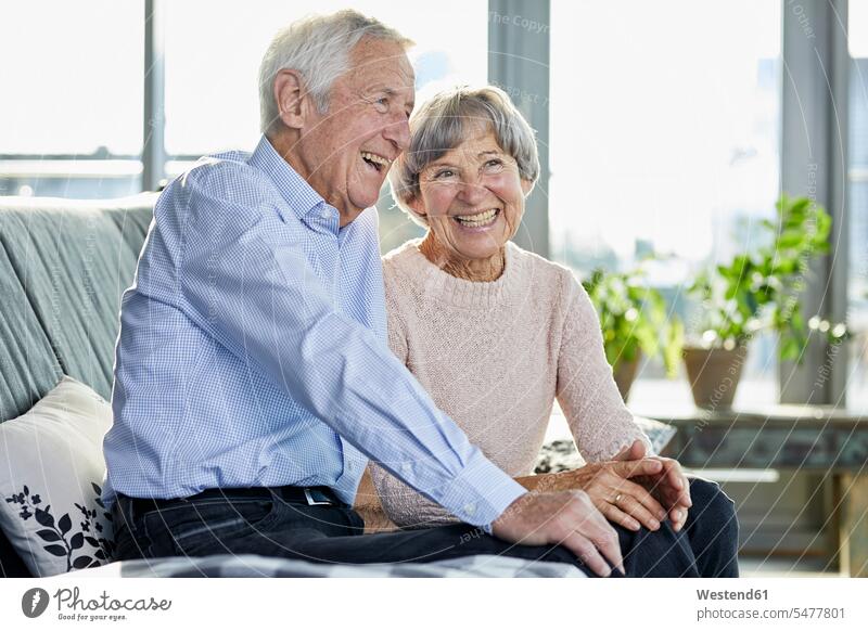 Portrait of laughing senior couple sitting together on couch settee sofa sofas couches settees elder couples senior couples twosomes partnership Laughter