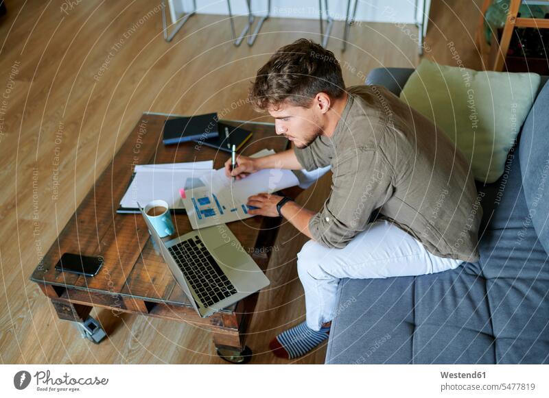 Male entrepreneur writing on paper while sitting with laptop at home color image colour image indoors indoor shot indoor shots interior interior view Interiors
