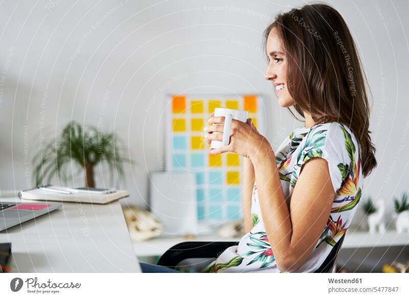 Smiling young woman drinking coffee at desk desks smiling smile females women Coffee Table Tables Adults grown-ups grownups adult people persons human being