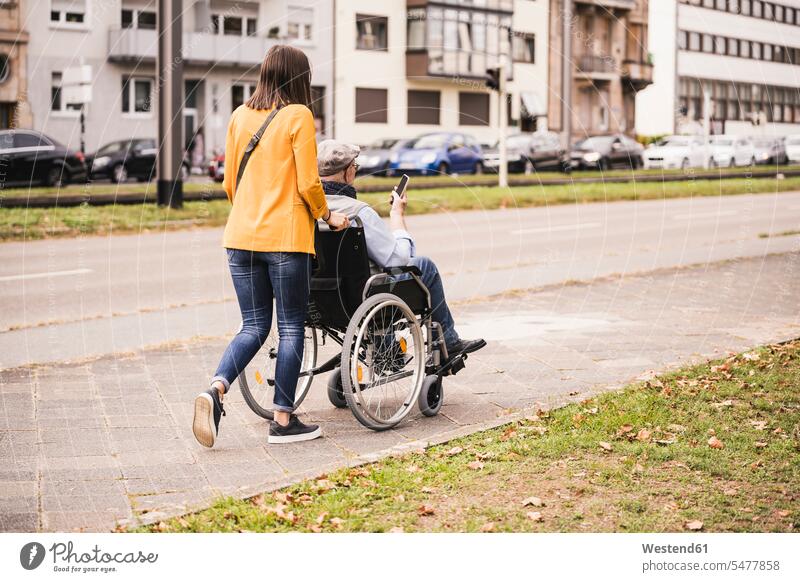 Back view of young woman pushing senior man with smartphone in wheelchair on pavement human human being human beings humans person persons caucasian appearance