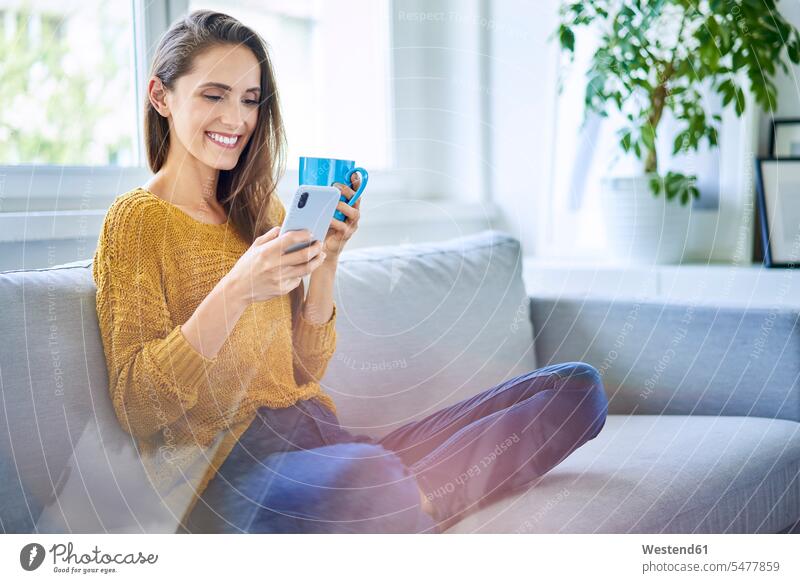 Smiling young woman sitting on sofa with smartphone and cup of coffee human human being human beings humans person persons celibate celibates singles