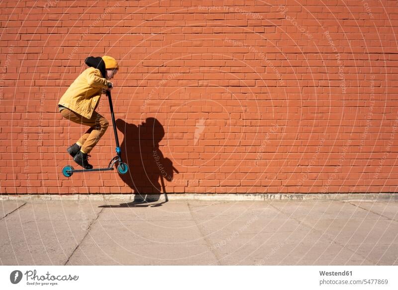 Boy performing stunt with push scooter on sidewalk against brick wall during sunny day color image colour image outdoors location shots outdoor shot