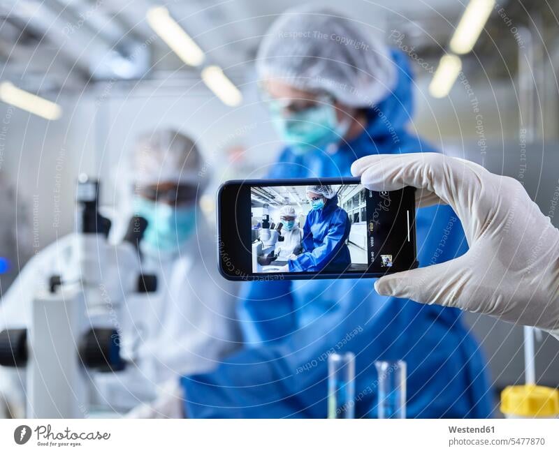 Hand holding smartphone, photographing chemists, working in industrial laboratory Smartphone iPhone Smartphones Chemical Laboratory Protective Suit At Work