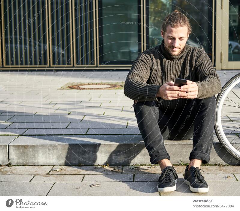 Portrait of bearded young man sitting on curb next to bicycle using cell phone beside men males Curbside kerb Smartphone iPhone Smartphones portrait portraits
