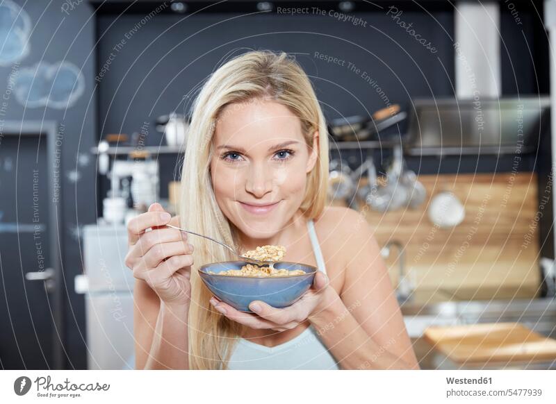 Portrait of smiling blond woman in the kitchen eating cereals domestic kitchen kitchens portrait portraits females women smile blond hair blonde hair Cereal