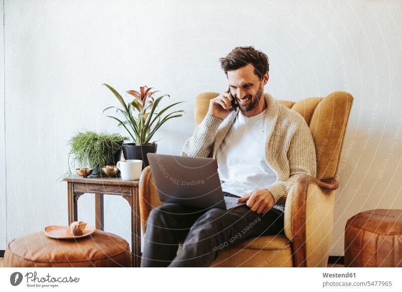 Smiling man on the phone sitting on armchair at home looking at laptop human human being human beings humans person persons caucasian appearance