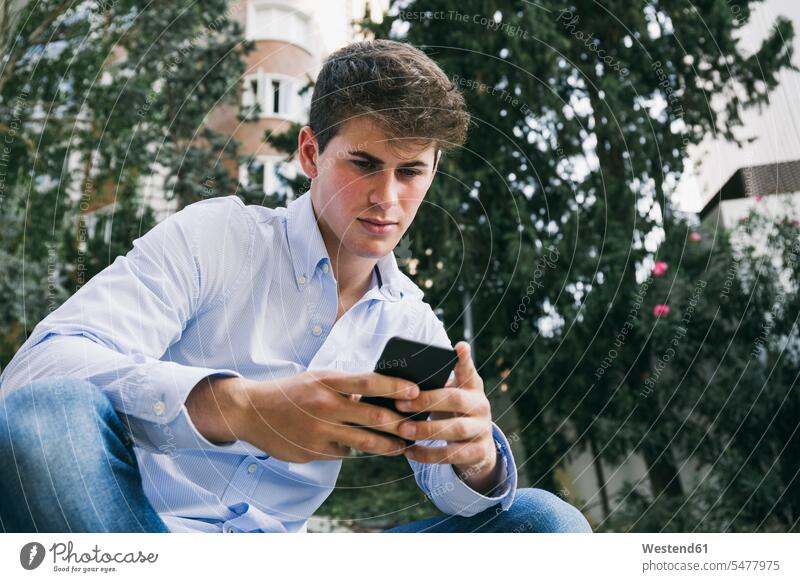 Handsome young man using smart phone while sitting against trees in city color image colour image Spain outdoors location shots outdoor shot outdoor shots day