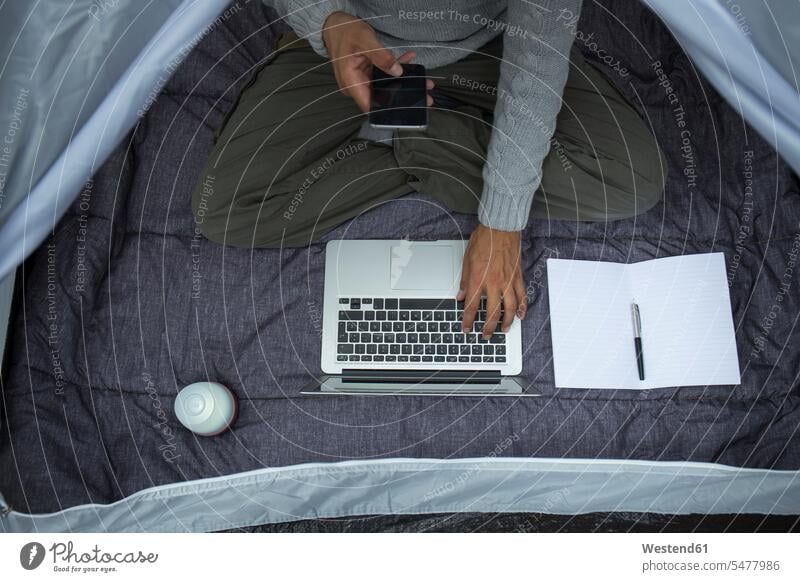Man sitting in tent using laptop and cell phone, partial view man men males Laptop Computers laptops notebook tents Smartphone iPhone Smartphones Seated use