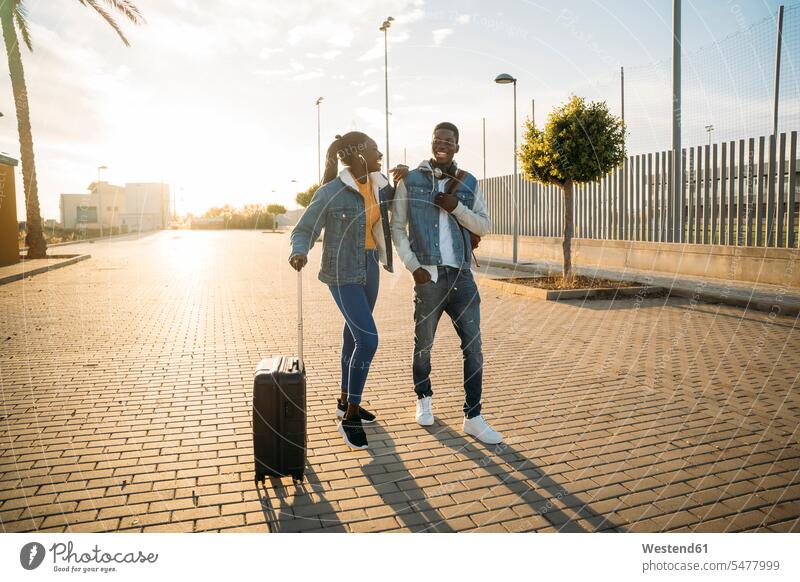 Happy heterosexual couple with wheeled luggage walking on street during sunny day color image colour image outdoors location shots outdoor shot outdoor shots