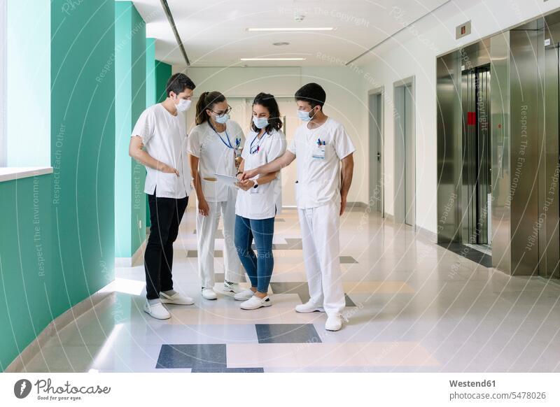 Doctors wearing surgical masks discussing over digital tablet while standing in corridor at hospital color image colour image Spain indoors indoor shot