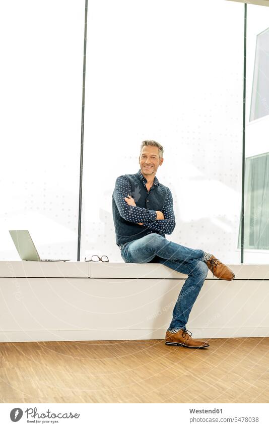 casual businessman sitting on windowsill in office building, with laptop next to him human human being human beings humans person persons caucasian appearance