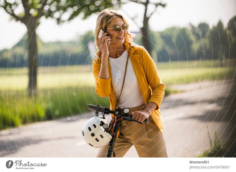 Happy mature woman using smart phone while standing with push scooter on roadside color image colour image outdoors location shots outdoor shot outdoor shots