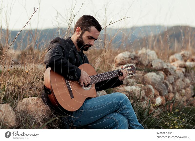 Male musician with beard playing the guitar in the countryside musicians guitars guitarist guitarists sitting Seated making music playing music make music