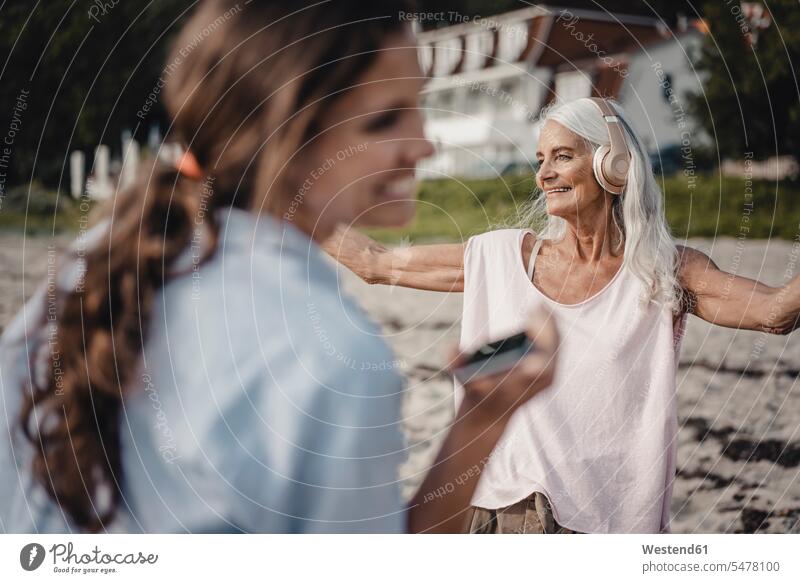 senior woman dancing on the beach, daughter laughing in foreground generation headphone headset telecommunication phones telephone telephones cell phone