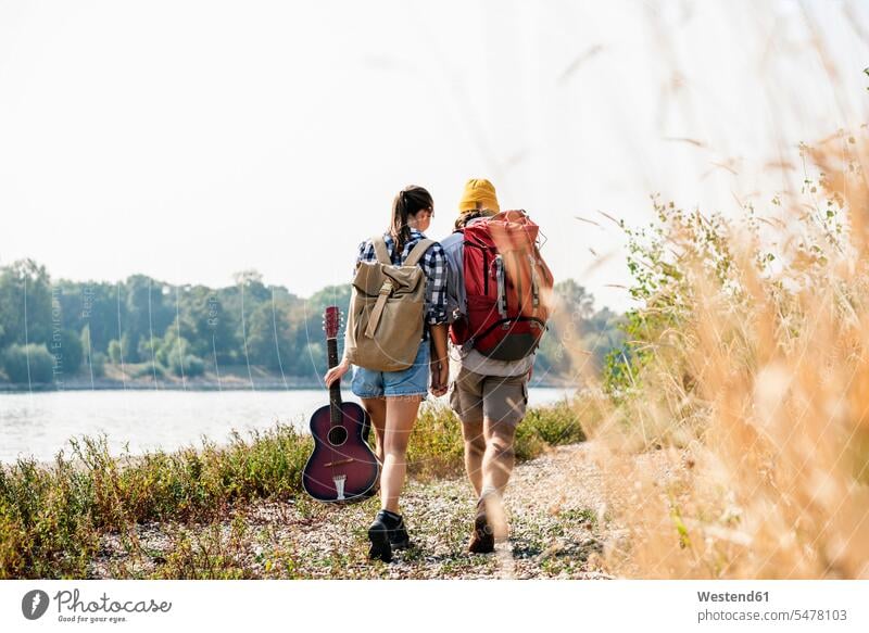 Rear view of young couple with backpacks and guitar walking at the riverside rucksacks back-packs guitars riverbank going River Rivers twosomes partnership