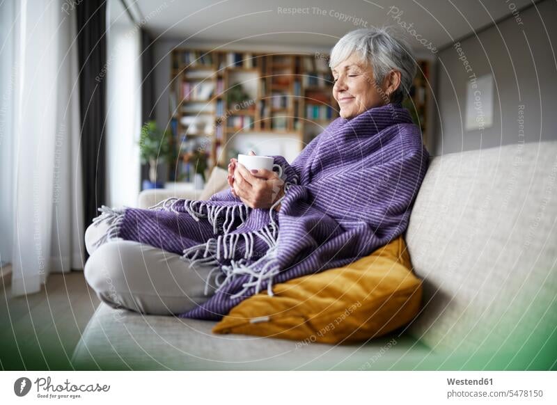 Thoughtful senior woman with blanket holding coffee cup while sitting on sofa at home color image colour image indoors indoor shot indoor shots interior