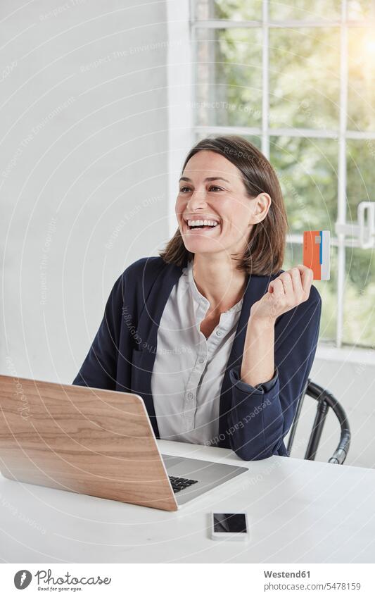 Laughing businesswoman using laptop on desk holding card Laptop Computers laptops notebook desks cards laughing Laughter businesswomen business woman