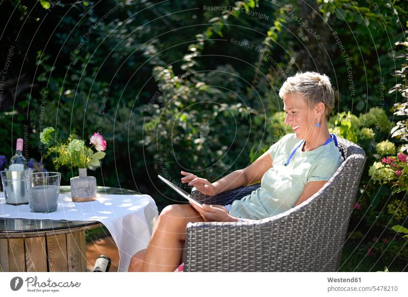 Smiling woman sitting in the garden using digital tablet smiling smile Seated females women gardens domestic garden digitizer Tablet Computer Tablet PC