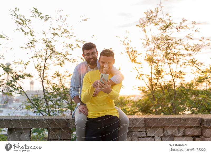 Gay couple using cell phone outdoors at sunset associate associates partner partners partnerships telecommunication phones telephone telephones cell phones