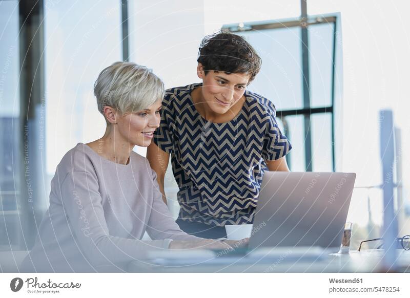 Two smiling businesswomen sharing laptop at desk in office smile share offices office room office rooms working At Work Female Colleague businesswoman
