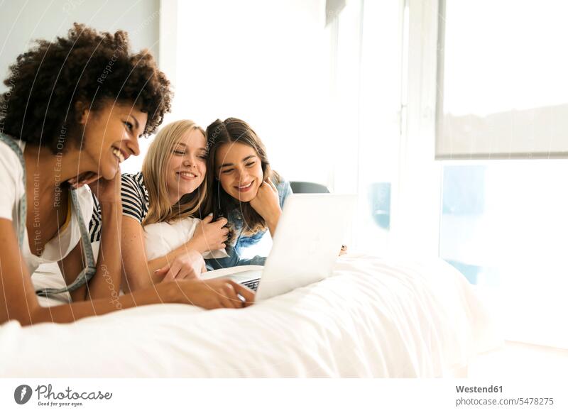 Three happy girlfriends lying on bed sharing laptop laying down lie lying down female friends beds Laptop Computers laptops notebook happiness share woman