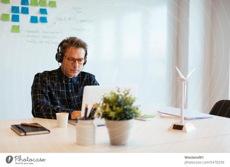 Businessman wearing headphones using laptop at desk in office Business man Businessmen Business men offices office room office rooms desks Laptop Computers