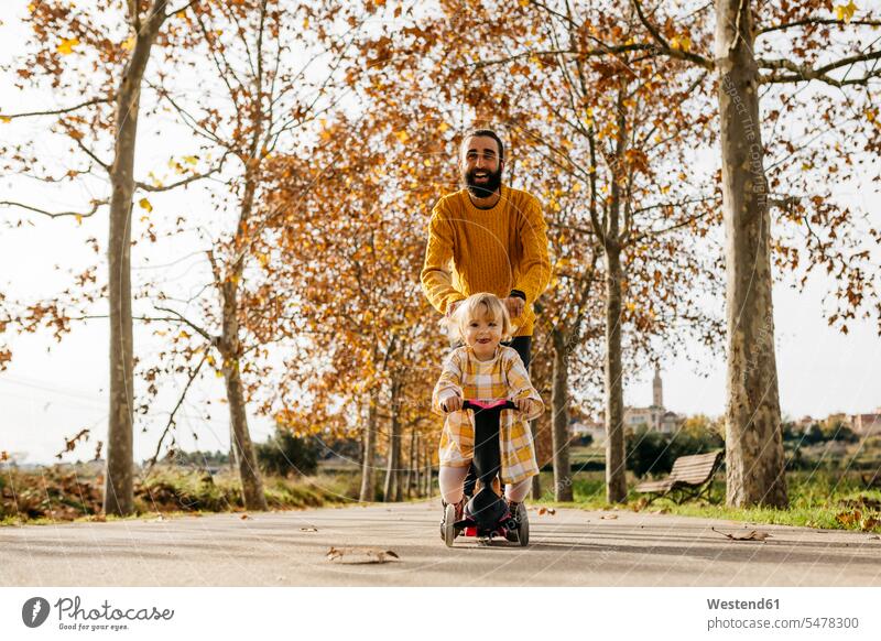 Father and daughter enjoying a morning day in the park in autumn, girl on scooter autumnal autumnally parks tricycle father fathers daddy dads papa daughters
