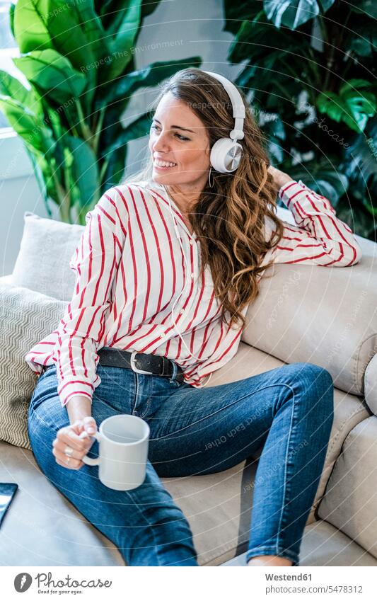Smiling woman with cup of coffee and headphones sitting on couch headset Coffee Cup Coffee Cups Seated females women settee sofa sofas couches settees Drink
