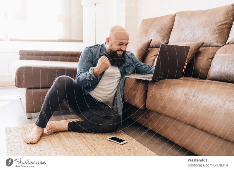 Happy businessman cheering with clenching fist while looking at laptop in living room color image colour image casual clothing casual wear leisure wear