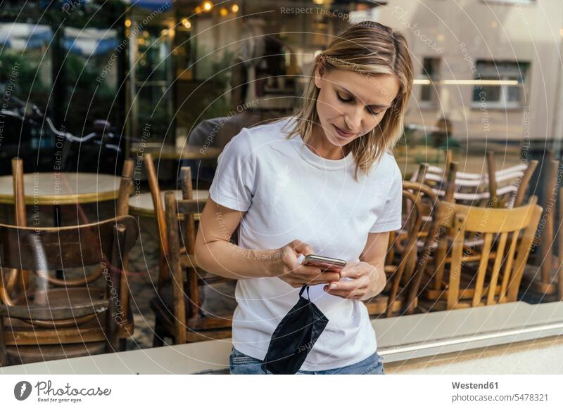 Portrait of woman with protective mask in her hand text messaging in front of a closed coffee shop human human being human beings humans person persons