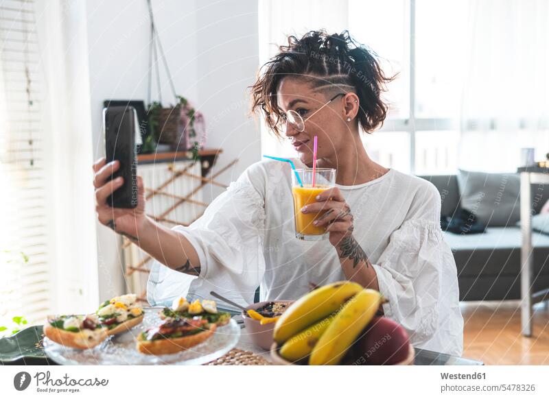 Portrait of young woman taking smart phone selfie while eating breakfast indoors indoor shot indoor shots interior interior view Interiors day daylight shot